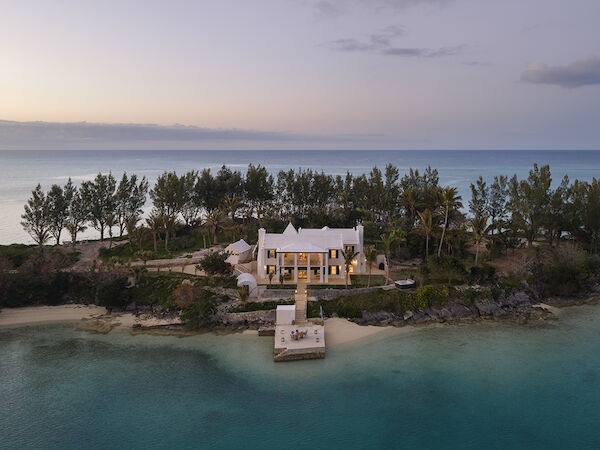 A white beach house sits on a rocky cape with a private dock, surrounded by trees, overlooking a turquoise bay at sunset.