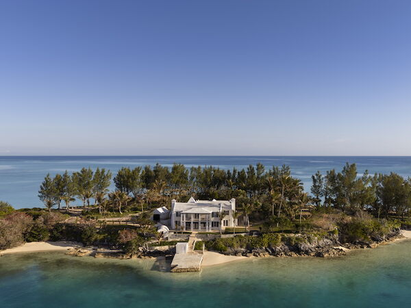 A white house sits on a small island with a stone jetty, surrounded by trees and clear blue water, under a bright sky.