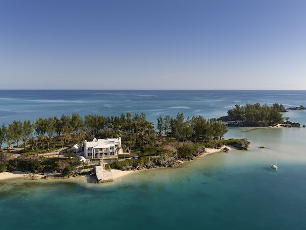 A small coastal house sits on a tree-lined peninsula, surrounded by turquoise water and a sandy beach, with a calm sea and clear blue sky.