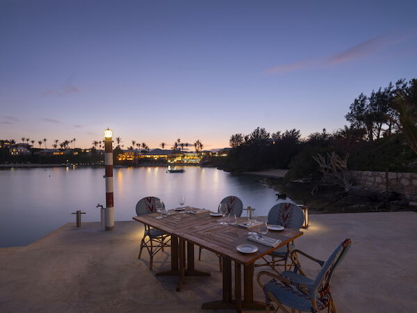A serene lakeside dinner setup at dusk with a wooden table, four chairs, and a calm water view reflecting asoft sunset glow.