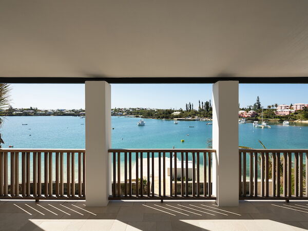 A seaside view from a covered balcony with a wooden railing, turquoise water, boats, and a distant shoreline under a sunny sky.