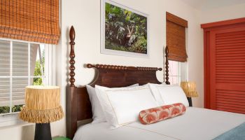 A cozy bedroom features a wooden bed with white linens, a decorative pillow, two bedside tables with lamps, and a framed artwork above the bed.