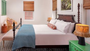 A cozy bedroom with a large bed, a blue blanket, decorative lamps, and an armchair. The room has a relaxing ambiance with wooden blinds.