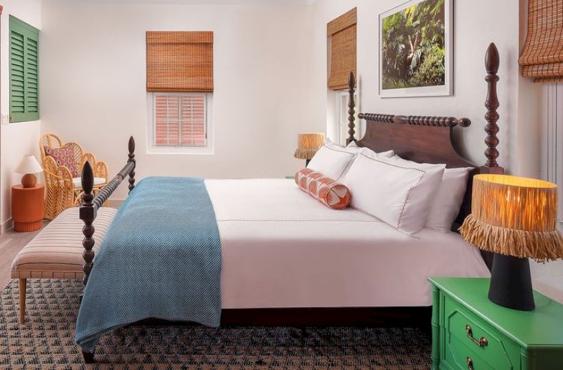 A cozy bedroom with a large bed, a blue blanket, decorative lamps, and an armchair. The room has a relaxing ambiance with wooden blinds.