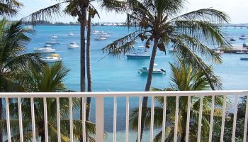 A view of a tropical seaside with palm trees, boats in the water, and a white railing in the foreground ending the sentence.