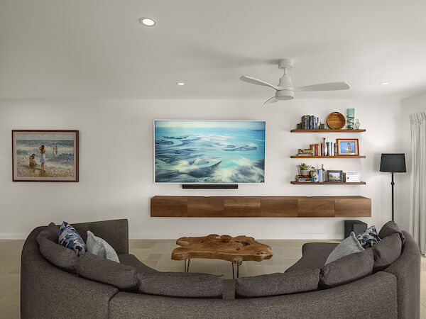 A bright, modern living room with a large curved sofa facing a wall-mounted TV, floating wooden shelf, and coastal decor on shelves, including a round coffee table and ceiling fan.