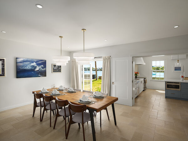 A bright, open dining area with a wooden table set for six, modern chairs, artwork on white walls, and a view into a sunlit kitchen beyond.