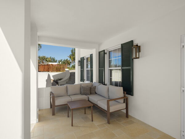 A bright covered patio with white walls, a beige sectional sofa, wooden coffee table, and a view of a fenced yard beyond the seating area.