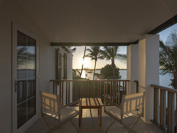 A cozy balcony with two chairs and a small table, overlooking palm trees and a sunset by the water.