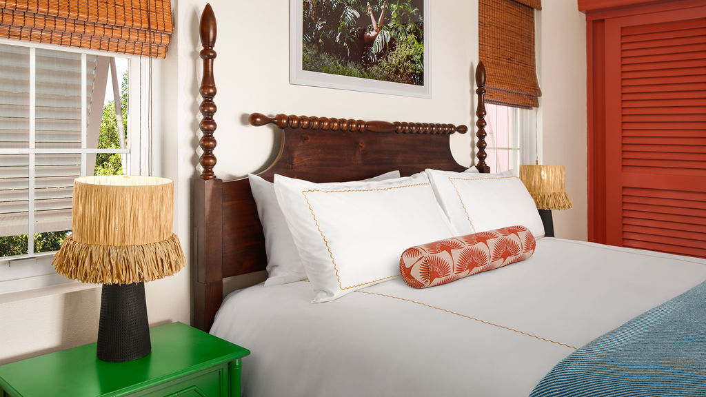 A cozy bedroom with a dark wooden bed frame, white bedding, a patterned bolster pillow, two bamboo-themed lamps, and a colorful picture above the bed.