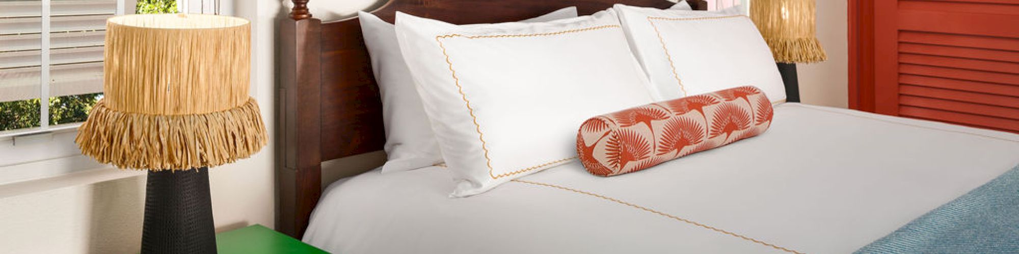 A cozy bedroom with a dark wooden bed frame, white bedding, a patterned bolster pillow, two bamboo-themed lamps, and a colorful picture above the bed.