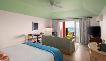 A modern hotel room with a view of the ocean, featuring a bed, a seating area, a TV, a table with chairs, and an open balcony door.