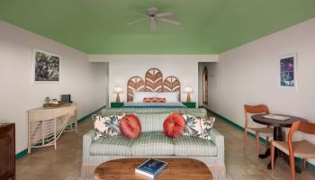 A cozy living room with green accents features a green sofa, two red and white pillows, a wooden coffee table, and a TV on a wicker stand.
