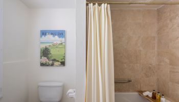 The image shows a clean bathroom divided into two sections: a toilet area with a Bermuda poster and a bathtub with a shower curtain and toiletries.