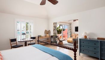 The image features a bright, airy bedroom with a bed, a ceiling fan, a dresser, and a small seating area near windows overlooking an outdoor space.
