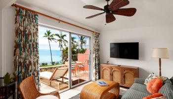 A cozy living room with tropical décor, featuring a sofa, wicker chair, TV, ceiling fan, and a sliding glass door leading to a balcony with an ocean view.