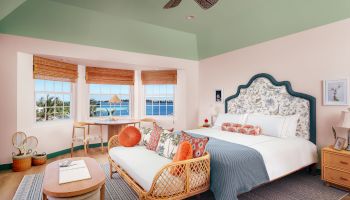 A cozy bedroom features a large bed with decorative pillows, a wicker sofa, a wooden desk by large windows, and a ceiling fan, bathed in natural light.