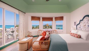 A brightly lit bedroom with a large bed, wicker furniture, and windows offering a view of the ocean, along with a ceiling fan and sliding glass doors.