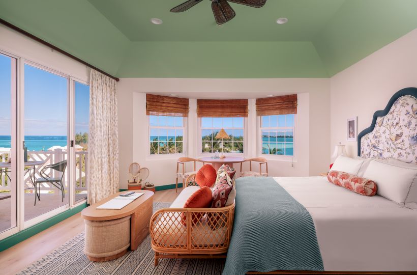 A brightly lit bedroom with a large bed, wicker furniture, and windows offering a view of the ocean, along with a ceiling fan and sliding glass doors.