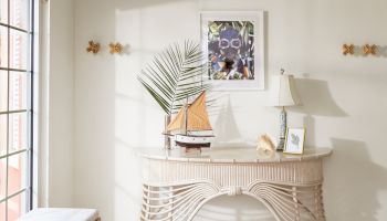 A decorative room corner featuring a wicker console table with a model sailboat, framed art above, a lamp, and beach-themed accents on the wall.