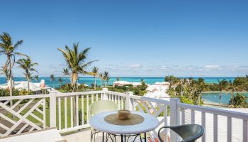 A picturesque balcony view with a table and two chairs, overlooking lush greenery, palm trees, and a serene ocean with a clear blue sky.