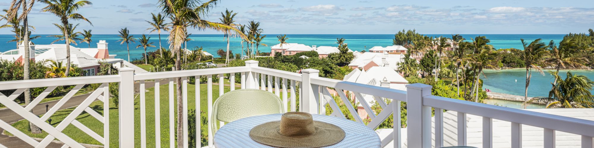 A scenic view featuring a small table with a hat, chairs on a balcony, overlooking palm trees, houses, and the ocean under a clear blue sky.