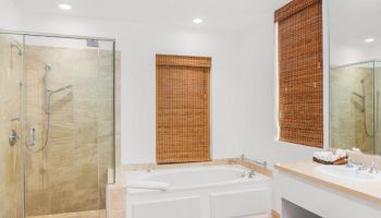 A bathroom with a glass-enclosed shower, bathtub, wooden blinds, countertop sink, and mirror. The setting is neat and well-lit.