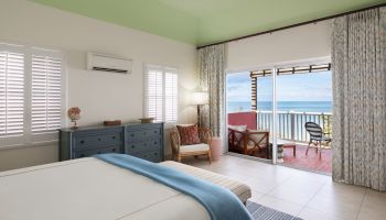 A bedroom with a bed, dresser, armchair, and open balcony with ocean views. The room is bright and airy, featuring pastel colors and natural light.