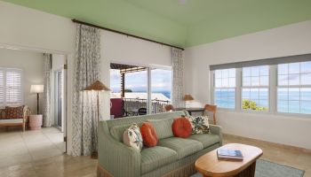 This image shows a bright, airy living room with ocean views, featuring a green ceiling, sofa, coffee table, and large windows.