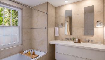The image shows a modern bathroom with a bathtub, dual sinks, two mirrors, and beige tile walls. It has a window, towels, and various toiletries.