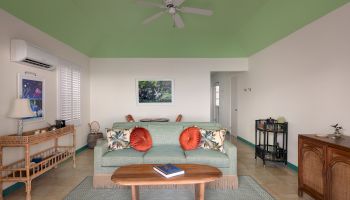 A cozy living room with a mint green ceiling, a sofa with orange pillows, a wooden coffee table, a ceiling fan, and various pieces of wooden furniture.