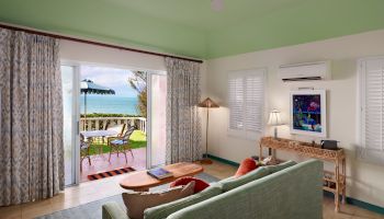 A cozy living room with a sea view, featuring a green sofa, TV, air conditioner, lamp, and sliding doors leading to a patio with a table and umbrella.