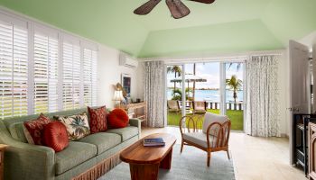 A cozy, tropical-themed living room with a green ceiling, wooden furniture, a sofa, glass doors to a patio, and a view of the ocean.
