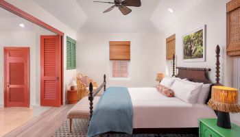 A cozy bedroom with a large bed, colorful accents, a ceiling fan, wicker chairs, and a vibrant green nightstand with table lamps.