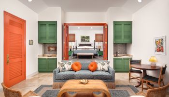 A modern, colorful living room features a sofa, armchairs, a wooden coffee table, and a small dining area, leading into a stylishly furnished bedroom.