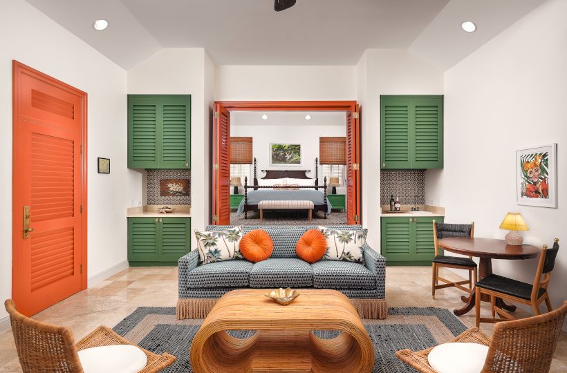 A modern, colorful living room features a sofa, armchairs, a wooden coffee table, and a small dining area, leading into a stylishly furnished bedroom.
