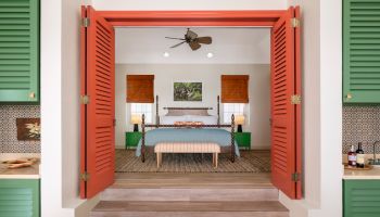 The image shows a cozy bedroom with green shutters, a bed with white linens, orange doors, a ceiling fan, and a bench at the foot of the bed.