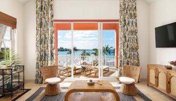 A stylish living room features wicker furniture, a modern TV, tropical curtains, and a glass door opening to a balcony with an ocean view, ending the sentence.