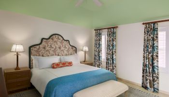 A bedroom with a large bed, floral-patterned headboard, blue blanket, and a bench at the foot. Two lamps on bedside tables and patterned curtains.