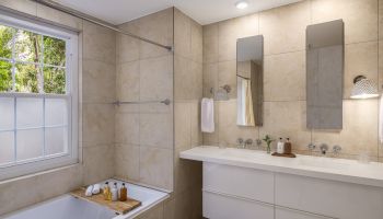 Modern bathroom with a double sink, two mirrors, a bathtub with a wooden tray holding toiletries, beige tile walls, and a window with natural light.