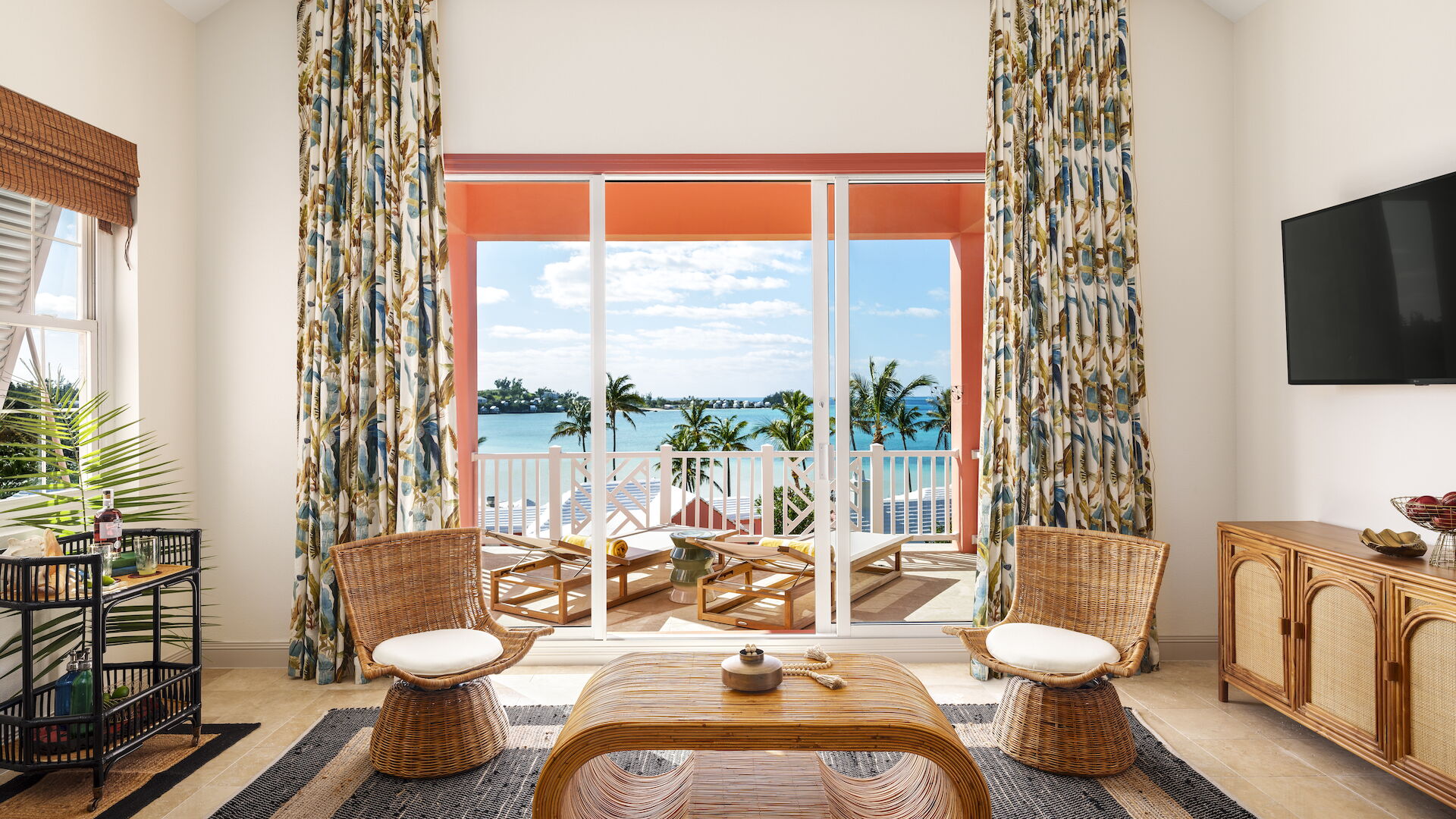 A cozy living room with wicker furniture, colorful curtains, and a view of a sunny balcony. The room features a TV and decorative plants.