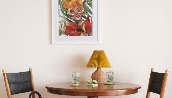 A small wooden table with a lamp, two glasses, and a decor item on it, flanked by two chairs. A floral painting hangs above the table.