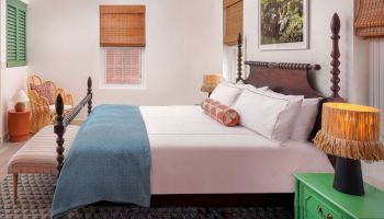 A neatly organized bedroom with a double bed, green side table, lamps, wicker chairs, and windows with blinds, exuding a cozy ambiance.