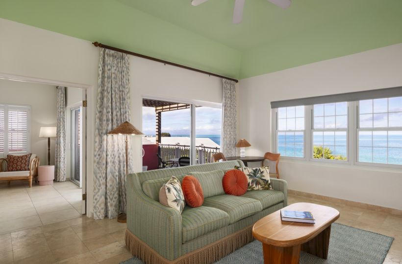 A living room with green sofa, orange cushions, coffee table, large windows with ocean view, ceiling fan, and doorway to another room, ending the sentence.