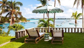 The image shows two lounge chairs under a parasol on a lawn overlooking a tropical beach with palm trees and boats in the background.