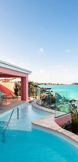 A pink seaside villa with a curved pool overlooking turquoise ocean; sunny sky, glass railing, and lush greenery in view.