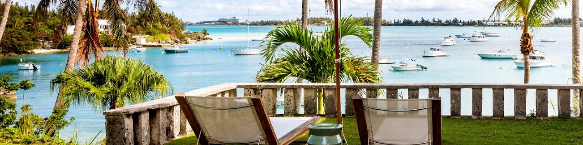 Two lounge chairs under a large umbrella overlook a scenic beach with palm trees, lush grass, clear blue water, and distant boats.