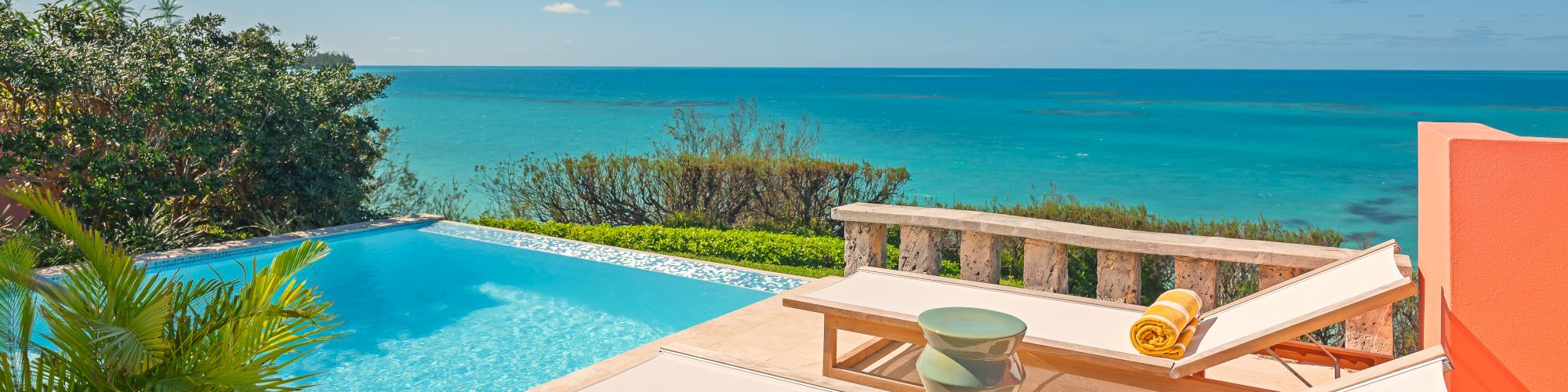 The image shows a coastal poolside scene with two lounge chairs, yellow towels, and a clear blue ocean view under a bright sky.
