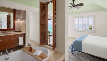 This image shows a bathroom with a bathtub and a view into a bedroom with a bed adorned with a blue throw. Both rooms have light, airy decor.