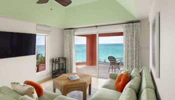 A living room with green furniture, a ceiling fan, and a view of the ocean through open sliding doors, leading to a patio with a table and chairs.